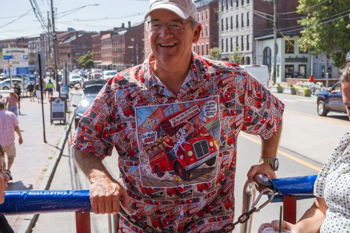 Man in colorful shirt on bus, smiling with people around, urban street in background.
