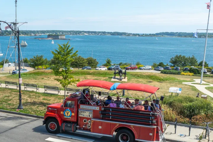 Red open-top tour bus by a waterfront, people onboard, sailboats in the distance.