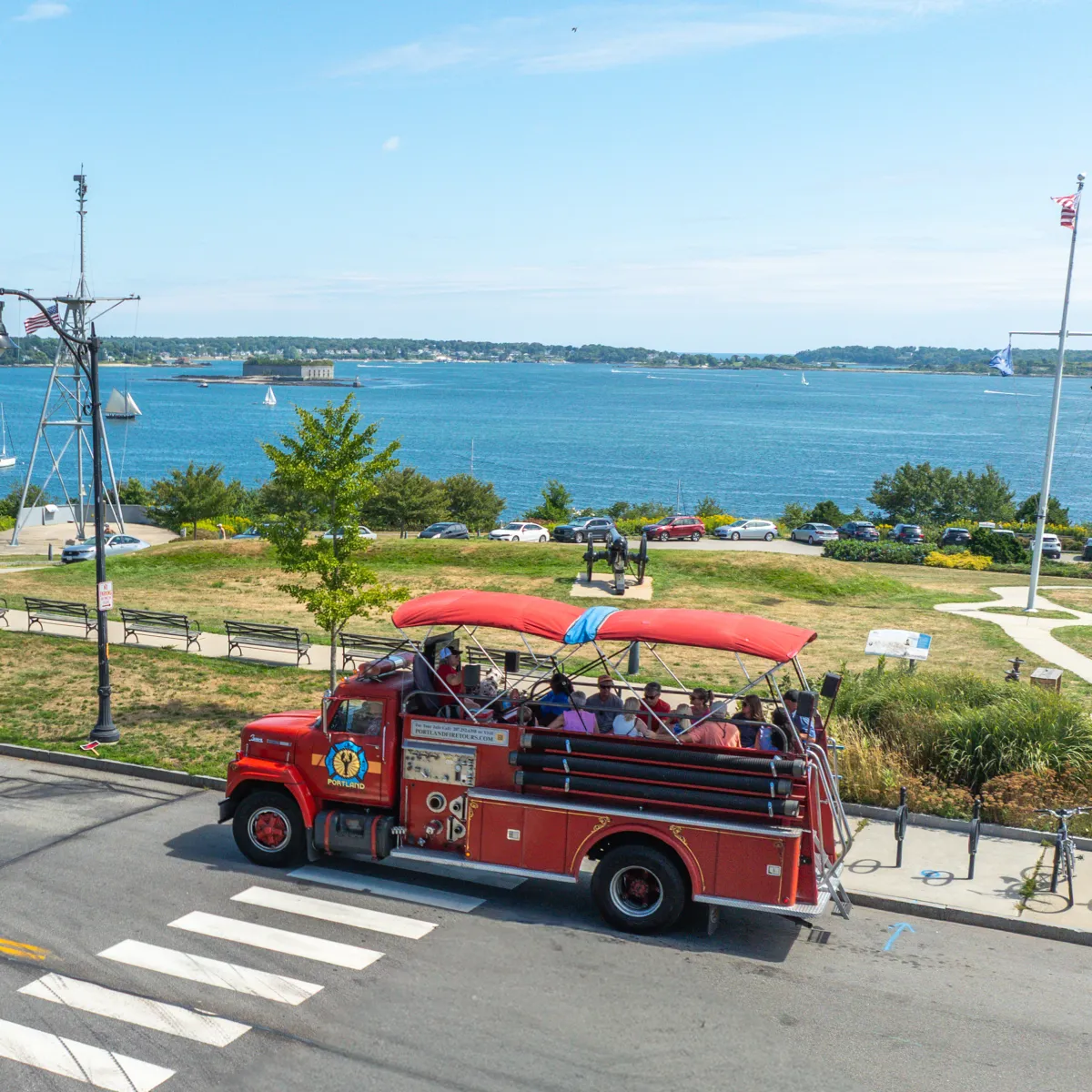 Red open-top tour bus by a waterfront, people onboard, sailboats in the distance.