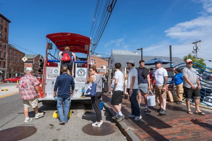 People lined up to board a tour bus on a sunny street.