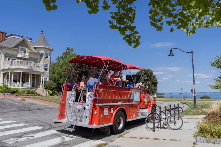 Fire truck tour vehicle with passengers near a historic house and seaside view.