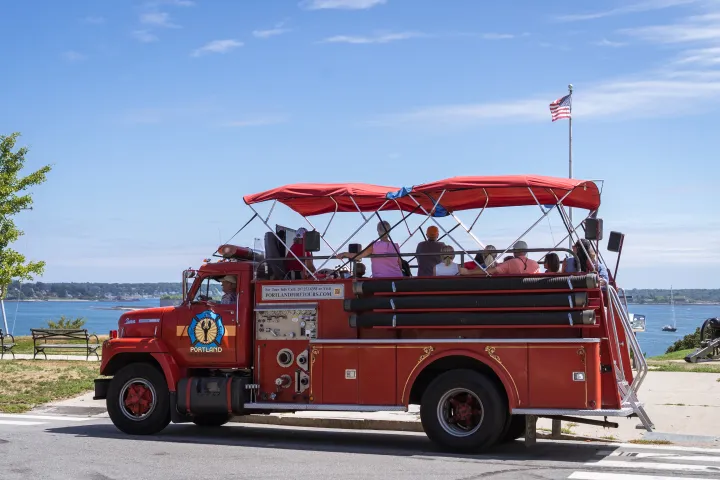 Red tour bus on street near waterfront, with people inside and American flag on top.